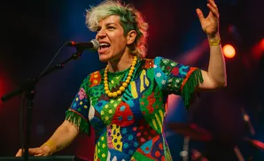 A person with green-tipped hair sings into a microphone at a concert, wearing a brightly colored, patterned dress and a yellow necklace.