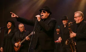 A band performs on stage with a lead singer, guitarist, and saxophonist in the foreground.