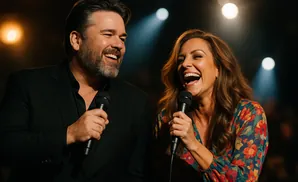 A man and woman sing into microphones, both with wide, joyful smiles, on a dimly lit stage.