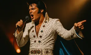 Elvis Presley impersonator in a white, bejeweled jumpsuit and blue cape, singing into a microphone.