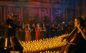 Conductor leads orchestra in a candlelit performance with a cellist in the foreground.