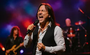 A male singer belts into a microphone on stage, with a guitarist and drummer blurred in the background.