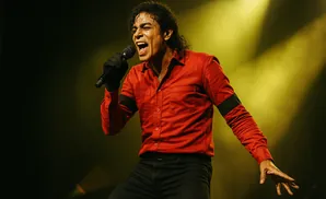 A man in a red shirt and black pants sings into a microphone on a dimly lit stage.
