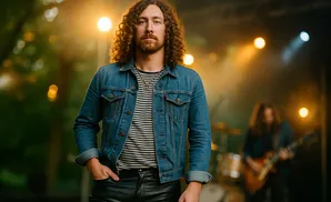 Man with curly hair in a denim jacket and leather pants on stage with a band.
