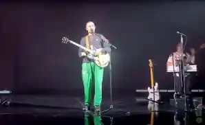 dermot kennedy performing on stage wearing black long sleeved shirt and green pants.