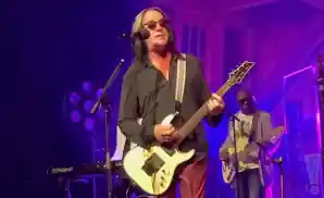 todd rundgren performing on stage playing the electric guitar with purple light behind him.