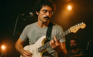 Man with curly hair, mustache, and glasses plays a white electric guitar on stage.