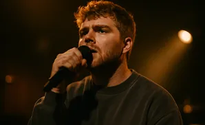 A man with reddish hair sings into a microphone in a dimly lit room.