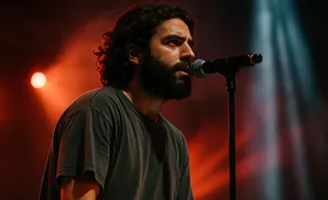 A man with dark, curly hair and a beard sings into a microphone on a dimly lit stage with red and blue spotlights.