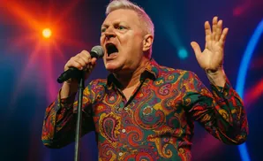 A man with white hair sings into a microphone, wearing a colorful paisley shirt and raising his hand on stage.