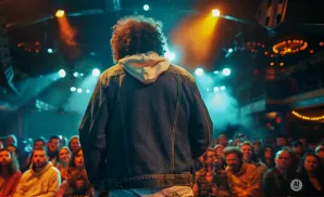 A performer with an afro stands on stage with their back to the audience, illuminated by blue and orange lights.