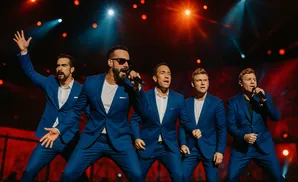 The Backstreet Boys perform in matching blue suits, singing on a stage with red and blue lighting.