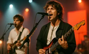 A male singer and guitarist performs on stage with a band, illuminated by stage lights.