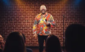 A comedian in a colorful shirt laughs on stage in front of a brick wall, holding a microphone.