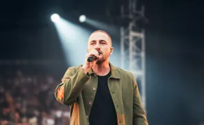 A man on stage holds a microphone and wears a green jacket with flower patches.