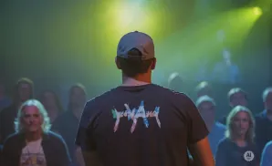 Man in a cap and t-shirt with a colorful design faces away from the camera, with an audience behind him lit by yellow spotlights.