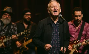 A man sings into a microphone, his eyes closed, while three guitarists play behind him.