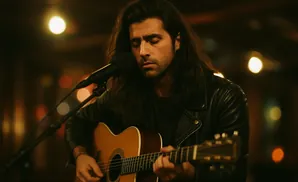Man with long hair playing an acoustic guitar and singing into a microphone.