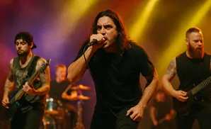 A rock band performs on stage with a lead singer at the microphone, a guitarist on the left, and another guitarist and drummer in the background.