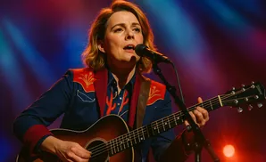 A woman in a blue and red western-style jacket plays an acoustic guitar and sings into a microphone.
