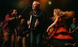 Man plays accordion on stage while a woman in a red dress dances and another man sings.