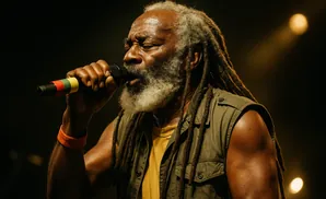 Man with dreadlocks and beard sings into a microphone, illuminated by stage lights.