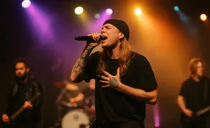 A male singer with blonde hair and tattoos performs on stage with a microphone and hand on his chest, under purple and orange lights.