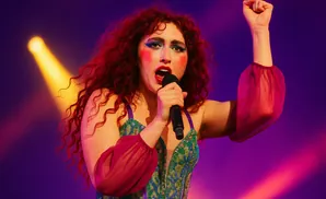 Singer with red curly hair, dramatic makeup, and a sparkly green outfit performs with a microphone, raising her fist.