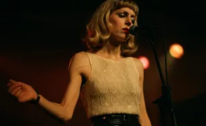 Singer with blonde hair and red lipstick performs into a microphone on a dark stage.