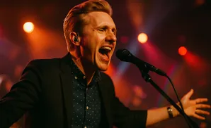 A man sings into a microphone on stage with red and orange lights behind him.