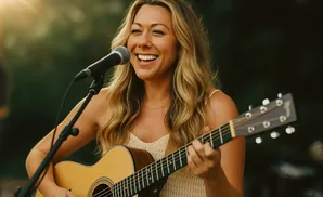 Woman with blonde hair playing an acoustic guitar and singing into a microphone.
