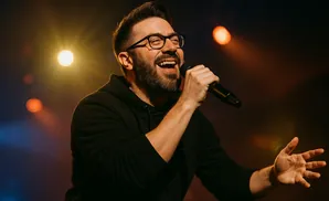 A man with glasses and a beard sings into a microphone, with warm stage lights behind him.