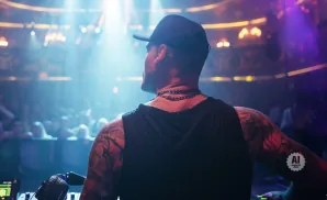 DJ wearing a black cap and tank top with tattoos, standing at a DJ booth with a crowd in the background.
