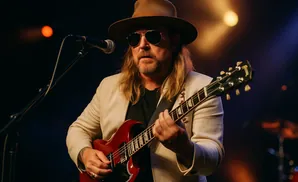 A man in a hat and sunglasses plays a red electric guitar on stage.