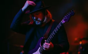 A man with a fedora and long hair plays a purple guitar on stage under colorful lights.