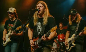 Band plays guitars on stage with singer in the middle.