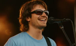 Musician wearing sunglasses and a blue shirt sings into a microphone.