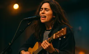 A woman with long, dark hair sings passionately into a microphone while playing an acoustic guitar.