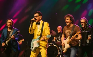 A band performs on stage with the lead singer in a yellow suit and a guitarist in a blue jacket.