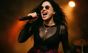 A female singer with long black hair, sunglasses, and a cross necklace sings into a microphone on stage.