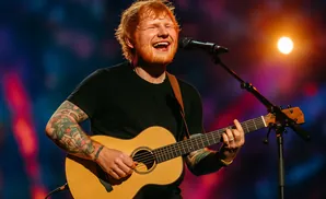 Ed Sheeran smiles while singing and playing an acoustic guitar on stage.