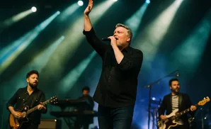 A lead singer performs with a microphone while bandmates play guitar and keyboards on a stage with bright lights.