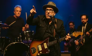 A man in a hat and glasses sings and plays a guitar on stage.