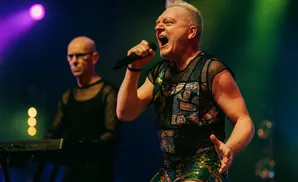 Singer performing on stage wearing a mesh top and sparkly shorts, with another band member in the background.
