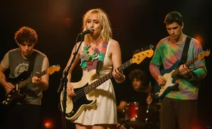 A band plays music on stage. The lead singer is in the middle, singing into a microphone and playing a bass guitar.