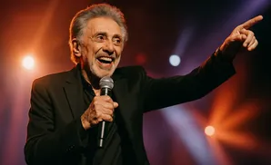 A charismatic older man with gray hair and a beard smiles warmly while performing on stage, holding a microphone and pointing toward the audience.