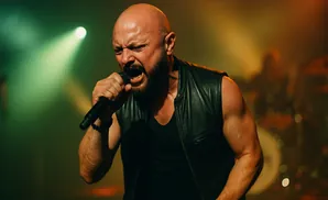 A bald, bearded man in a black leather vest sings intensely into a microphone on a stage with green and yellow lighting.