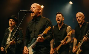 A bald man with a beard sings into a microphone while playing a guitar. Three other men play guitars behind him.