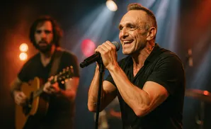 Singer on stage with microphone, guitarist in background.