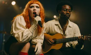 Woman with orange hair singing into a microphone while a man plays guitar.